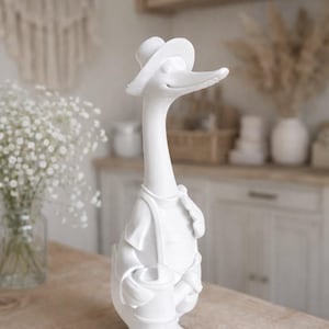 Puede incluir: Figura de pato de cerámica blanca con sombrero y peto, sosteniendo un pequeño cubo. El pato tiene un cuello largo y patas palmeadas. La figura está sobre una mesa de madera.