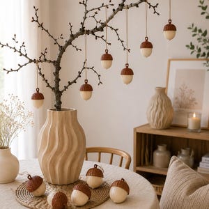 Könnte beinhalten: Eine Sammlung von eichelförmigen Ornamenten mit braunen, strukturierten Kappen und glatten, cremefarbenen Unterteilen. Mehrere Ornamente hängen an einem kahlen Ast in einer gewellten beigen Vase, während andere auf einer runden Flechtmatte liegen.