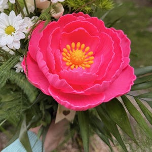 May include: A vibrant bouquet featuring a large, bright pink peony with a yellow center, surrounded by white daisies, a white rose, and green foliage. The arrangement is held together with a small tag.