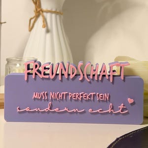 Könnte beinhalten: Ein lila rechteckiges Schild mit dem Wort "FREUNDSCHAFT" in rosa 3D-Schrift. Darunter steht "MUSS NICHT PERFEKT SEIN sondern echt" in rosa Schrift. Im Hintergrund eine weiße Vase mit Blumen.