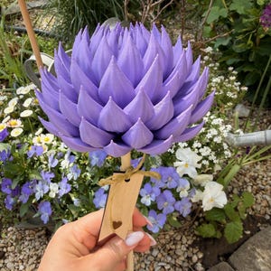 Könnte beinhalten: Eine große, lavendelfarbene, dekorative Blume auf einem Holzpflock. Die Blume hat viele spitze Blütenblätter. Ein kleines Holzschild mit einem Herz-Ausschnitt ist am Pflock befestigt. Die Blume wird vor einem Garten mit verschiedenen Blumen gehalten.