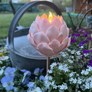 Puede incluir: Una lámpara solar de jardín en forma de loto rosa con un LED cálido y parpadeante. La luz está montada en una estaca de madera y rodeada de flores coloridas, incluyendo pensamientos azules y margaritas blancas. Una regadera de metal está en el fondo.