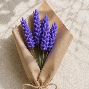 Op de afbeelding: Een klein boeket van vier paarse lavendelstelen verpakt in bruin kraftpapier. De stelen zijn samengebonden met een strik van dun, natuurkleurig jutekoord. De lavendelbloemen hebben een getextureerd, kraalachtig uiterlijk op dunne groene stelen.