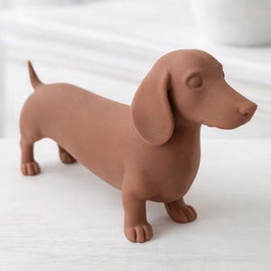 Könnte beinhalten: Eine braune Dackel-Figur steht auf einer weißen Oberfläche. Die Hundestatue hat einen langen Körper, kurze Beine und Schlappohren. Im Hintergrund befindet sich eine weiße Vase mit einem Strauß weißer, rosa und gelber Blumen.