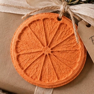 May include: A gift wrapped in brown paper with a cream-colored ribbon, featuring a textured orange citrus slice ornament, a sprig of pine, small white flowers, and a brown paper tag with the printed text Für dich and a small heart symbol.