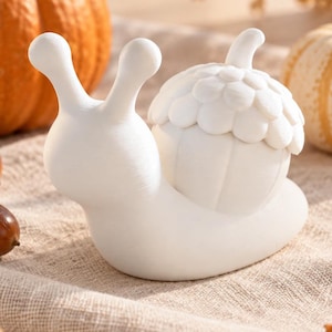 Könnte beinhalten: Eine mattweiße Schneckenfigur mit einem eichelförmigen Gehäuse und einer strukturierten Kappe. Die Schnecke hat zwei aufrechte Fühler und einen glatten, abgerundeten Körper. Sie steht auf einer strukturierten Stoffoberfläche neben herbstlicher Dekoration.