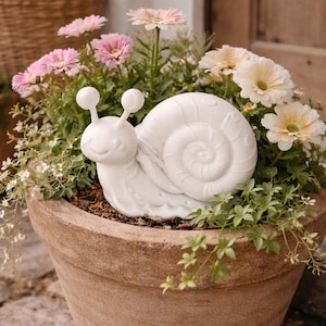 May include: A terracotta plant pot filled with pink and cream gerbera daisies and greenery. A white ceramic snail figurine sits amongst the flowers. Other wicker baskets with plants are in the background.
