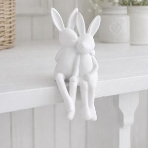 May include: A white ceramic figurine of two rabbits embracing. The rabbits are sitting with their legs dangling, one rabbit is kissing the other on the cheek. The background includes a white shelf, a wicker basket, and white flowers in jars.