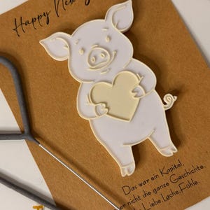 Könnte beinhalten: Eine Neujahrskarte mit einem weißen Schwein, das ein Herz hält, umgeben von Sektgläsern, Konfetti und einem Wunderkerzen. Die Karte lautet "Happy New Year!" und enthält ein deutsches Zitat.