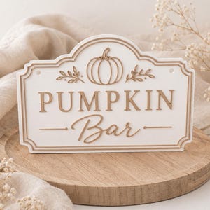 Könnte beinhalten: Ein rechteckiges weißes Schild mit gewellter Oberkante und der Aufschrift PUMPKIN Bar in erhabenen beigen Buchstaben. Das Design zeigt ein zentrales Kürbis-Symbol, flankiert von zwei kleinen Blattzweigen, alles in einem passenden Beigeton umrandet.