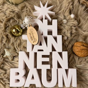 Könnte beinhalten: Eine weiße, dreidimensionale Weihnachtsbaumdekoration mit der Aufschrift "OH TANNENBAUM" und einem Stern oben. Ein Holzanhänger mit Text hängt an einer goldenen Kugel. Ein goldener Stern und eine Walnuss sind ebenfalls vorhanden.