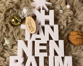 Puede incluir: Decoración navideña tridimensional blanca con la inscripción "OH TANNENBAUM" y una estrella en la parte superior. Una etiqueta de madera con texto cuelga de un adorno dorado. También hay una estrella dorada y una nuez.