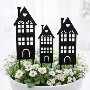 Może przedstawiać: Trzy czarne metalowe paliki ogrodowe w kształcie domu z wycięciami w kształcie serca i łukowatymi podstawami umieszczone są w białej doniczce wypełnionej białymi kwiatami stokrotki. Paliki mają detale komina i okna.