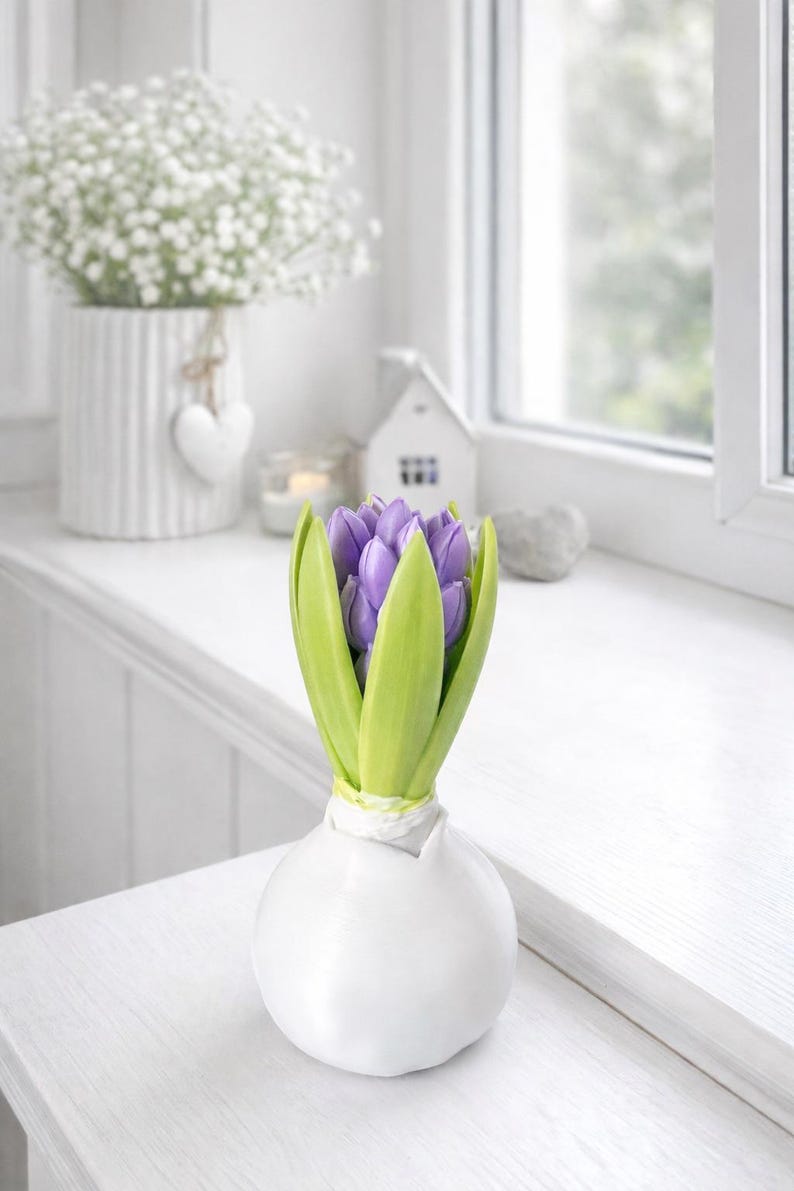 Puede incluir: Un jarr&oacute;n blanco en forma de bulbo contiene una flor de jacinto morada con hojas verdes. El jarr&oacute;n est&aacute; sobre una superficie blanca cerca de una ventana. Un peque&ntilde;o jarr&oacute;n blanco con flores blancas est&aacute; en el fondo.