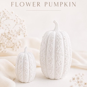 Op de afbeelding: Twee witte decoratieve pompoenen met ingewikkelde reliëfbloempatronen op hun oppervlak. De tekst FLOWER PUMPKIN is in een lichtbruin lettertype boven de pompoenen gedrukt, vergezeld van een kleine lijntekening van een bloem.