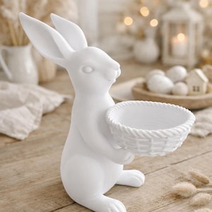 Può includere: Statuetta di coniglio in ceramica bianca che tiene un piccolo cestino intrecciato. Il coniglio ha lunghe orecchie ed è in posizione seduta. La statuetta è su una superficie di legno, con uno sfondo sfocato di elementi decorativi.