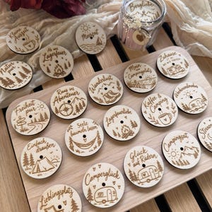May include: A collection of circular wooden tokens featuring laser-engraved nature and camping-themed illustrations and German text, arranged on a wooden board alongside a glass jar containing small pink items and a dried flower accent.
