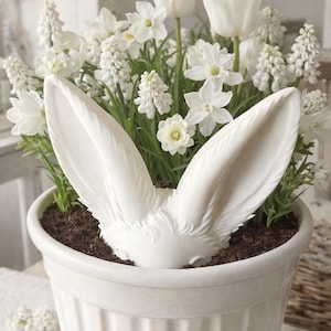 Könnte beinhalten: Ein weißer Keramiktopf gefüllt mit weißen Blumen und Grün. Eine dekorative weiße Hasenohren-Skulptur befindet sich in der Erde. Der Topf ist rund mit vertikalen Rillen.