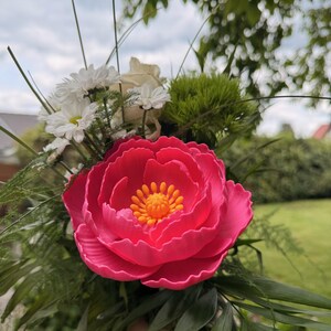 May include: A vibrant bouquet featuring a large, bright pink peony as the focal point. The peony has a detailed, layered petal design with a yellow center. Accompanying the peony are white daisies, a white rose, and green foliage, creating a colorful floral arrangement.