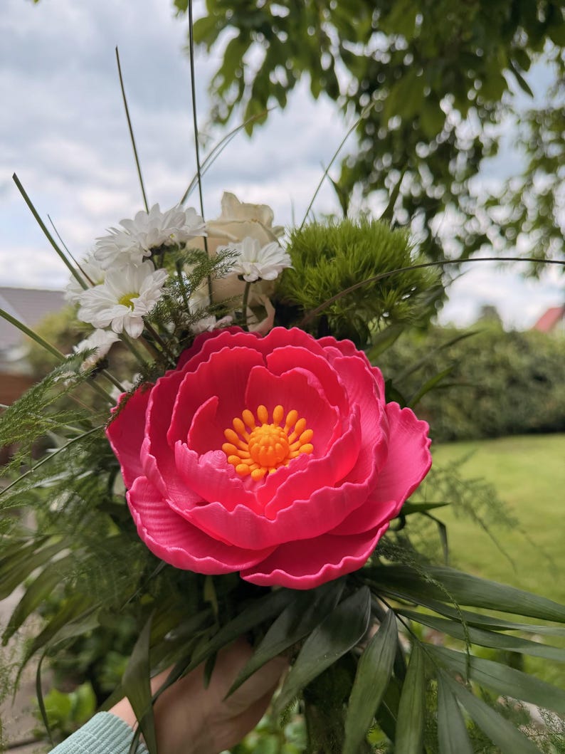 May include: A vibrant bouquet featuring a large, bright pink peony as the focal point. The peony has a detailed, layered petal design with a yellow center. Accompanying the peony are white daisies, a white rose, and green foliage, creating a colorful floral arrangement.