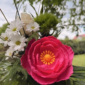 May include: A vibrant bouquet featuring a large, bright pink peony with a yellow center. The arrangement includes white daisies, a white rose, and green foliage. The flowers are set against a blurred background of greenery.