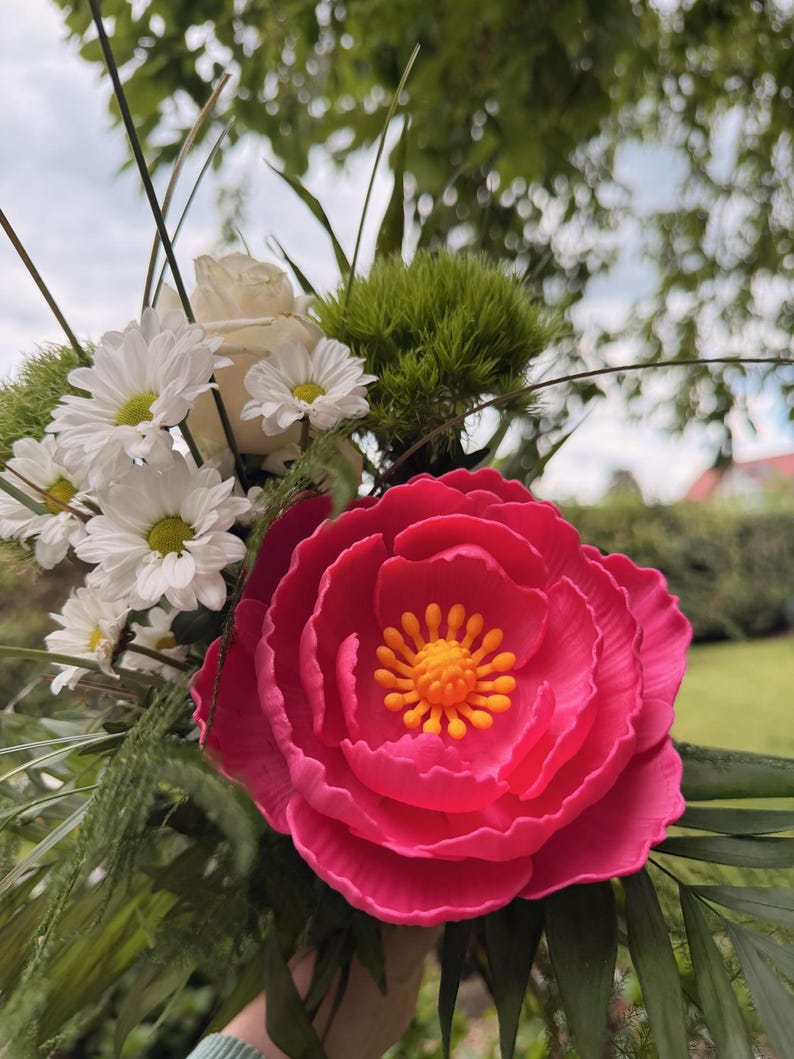 May include: A vibrant bouquet featuring a large, bright pink peony with a yellow center. The arrangement includes white daisies, a white rose, and green foliage. The flowers are set against a blurred background of greenery.