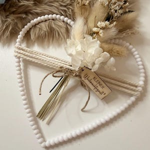 Könnte beinhalten: Herzförmiger Wandschmuck mit weißem Perlenrahmen. Die Mitte ist mit Trockenblumen und einem kleinen Holzanhänger verziert. Der Anhänger hat Text. Das Design integriert natürliche Elemente und neutrale Töne und schafft eine rustikale Ästhetik.