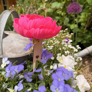May include: A vibrant pink, artificial peony flower on a wooden stake with a heart cutout, placed among purple and white pansies. A metal watering can is in the background, with purple allium flowers and greenery. A charming garden scene.