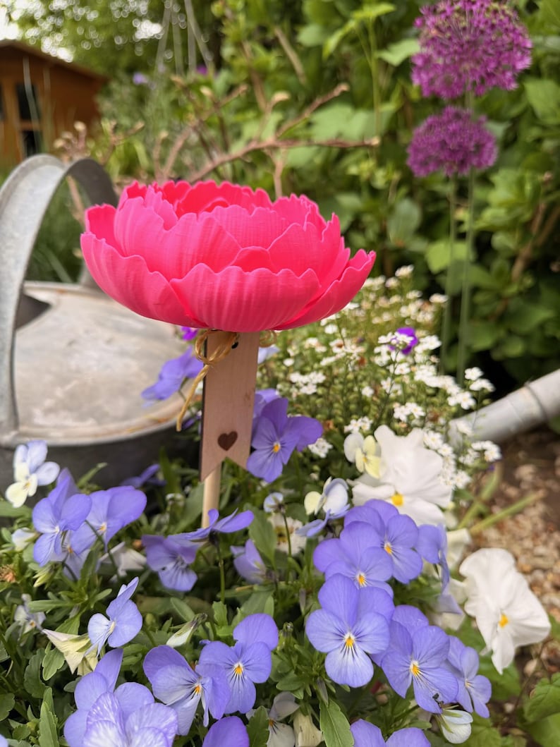 May include: A vibrant pink, artificial peony flower on a wooden stake with a heart cutout, placed among purple and white pansies. A metal watering can is in the background, with purple allium flowers and greenery. A charming garden scene.