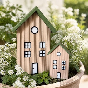 May include: Two wooden miniature houses with green roofs and light brown siding. The houses have black-framed windows and white doors. They are surrounded by white flowers and greenery in a terracotta pot.