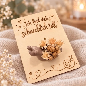 Op de afbeelding: Een lichtbruin slakvormig figuurtje met een bosje gedroogde beige bloemen op de rug, bevestigd op een rechthoekige houten kaart. De kaart bevat de lasergegraveerde Duitse tekst 'Ich find dich schnecklich toll', kleine hartjes en een slakillustratie.