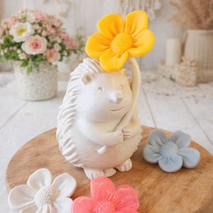 Puede incluir: Una figura de erizo de cerámica blanca sosteniendo una flor amarilla, con tres decoraciones en forma de flor en blanco, rosa y azul. Los artículos están dispuestos sobre una tabla de madera, sugiriendo un tema decorativo.