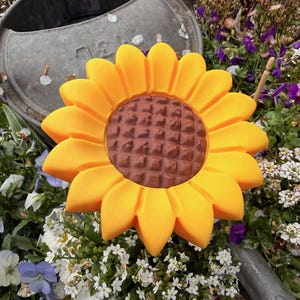 May include: A large, yellow sunflower garden ornament with a brown center. The sunflower is surrounded by white and purple flowers, with a galvanized metal watering can in the background. A decorative piece for the garden.