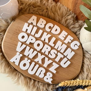 Op de afbeelding: Witte hoofdletter alfabetletters gerangschikt op een houten plank. De letters zijn in een schreefloze lettertype. De plank ligt op een zacht, lichtbruin oppervlak. Decoratieve elementen zijn een witte mok en kleine planten.