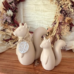 Puede incluir: Dos figuras de ardilla de color beige claro con superficies lisas y colas curvas. La ardilla más grande lleva un cordel de yute fino alrededor del cuello con una pequeña etiqueta circular que muestra una ilustración de una nuez blanca.