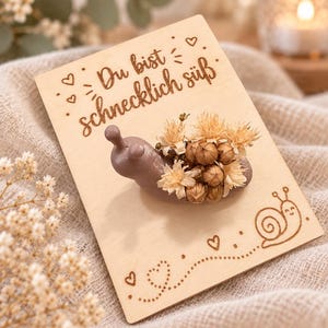 May include: A light brown snail figurine adorned with dried beige flowers, mounted on a rectangular wooden card. The card features the laser-engraved German text 'Du bist schnecklich süß', small heart icons, and a line drawing of a smiling snail.