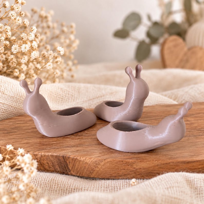 Op de afbeelding: Drie kleine, gladde, mauvekleurige slakvormige beeldjes, elk met een afgerond lichaam met twee rechtopstaande voelsprieten en een cirkelvormige inkeping op de rug, gerangschikt in een groep op een houten oppervlak.