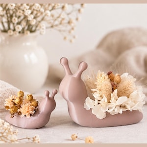 Op de afbeelding: Twee gladde, mauvekleurige plantenbakjes in de vorm van een slak in verschillende maten. Elke slak heeft een holle schelp die is gevuld met een arrangement van gedroogde bloemen, waaronder witte hortensiablaadjes en pluizige beige grassen.