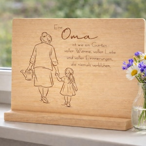 Könnte beinhalten: Holztafel mit graviertem Text und einer Zeichnung einer Großmutter und eines Kindes. Der Text lautet "Eine Oma ist wie ein Garten..." Rechts steht eine Blumenvase.