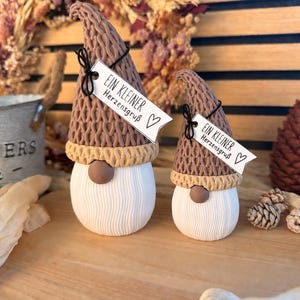 Puede incluir: Dos figuras de gnomos con sombreros texturizados de aspecto tejido marrón, bordes color canela y cuerpos blancos acanalados. Cada gnomo tiene una nariz redonda marrón y una etiqueta de papel blanco atada con hilo negro que dice "EIN KLEINER Herzensgruß" con un símbolo de corazón.