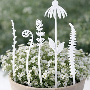 May include: A terracotta pot filled with small white flowers, accented by white, decorative plant stakes in various floral and botanical shapes. The stakes include a daisy, fern, and other stylized plant forms.