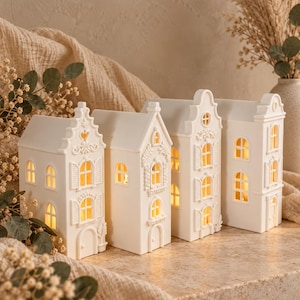 May include: A set of four white ceramic house-shaped lanterns with intricate architectural details including arched windows, shutters, and decorative gables. Each structure features cut-out openings that emit a warm, soft yellow glow from within.