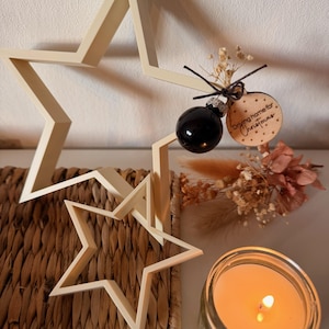 Könnte beinhalten: Eine weihnachtliche Dekoration mit zwei Holzsternen, einer schwarzen Kugel und einer brennenden Kerze in einem Glas. Ein Holzanhänger mit dem Text "Driving home for Christmas" ist ebenfalls zu sehen.