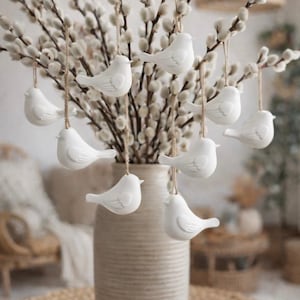 Könnte beinhalten: Eine dekorative Anordnung mit einer beigen Vase, gefüllt mit Weidenkätzchenzweigen. Weiße Keramik-Vogelornamente hängen an den Zweigen. Die Vase steht auf einer gewebten Matte.