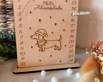 Può includere: Calendario dell'Avvento in legno con un cane bassotto con cappello da Babbo Natale e ghirlanda. Il calendario ha i numeri 1-24, il testo "MEIN Adventskalender" e disegni di fiocchi di neve. È attaccata una piccola clip bianca.
