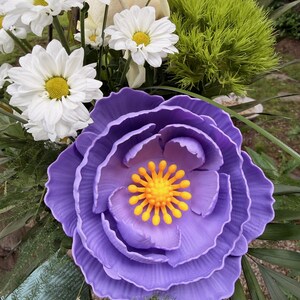 May include: A vibrant floral arrangement featuring a large, layered purple flower with a yellow center. White daisies and a cream-colored rose are also visible, along with green foliage. The arrangement is a mix of textures and colors.