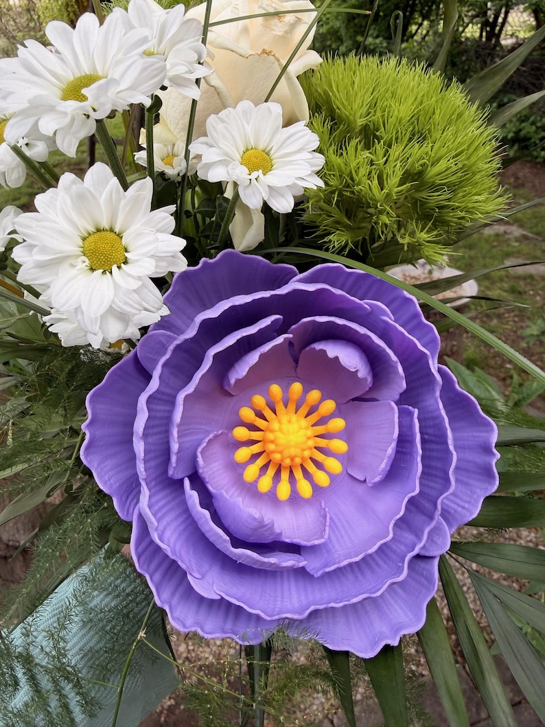May include: A vibrant floral arrangement featuring a large, layered purple flower with a yellow center. White daisies and a cream-colored rose are also visible, along with green foliage. The arrangement is a mix of textures and colors.
