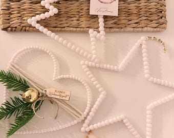 Puede incluir: Una composición navideña festiva con decoraciones en forma de estrella y corazón hechas de cuentas blancas. El arreglo incluye un pequeño ramo de flores secas, una vela en un portavelas de vidrio y una etiqueta con una ilustración de un caballito balancín y la frase "Merry Christmas".