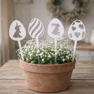 May include: A terracotta pot filled with white flowers and four white Easter egg-shaped decorations. Each egg has a different cutout design: a bunny, stripes, another bunny, and hearts. The pot sits on a wooden table.