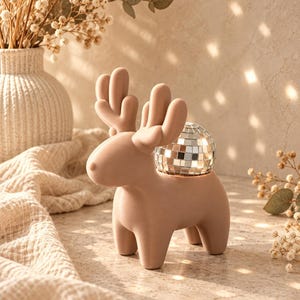 Puede incluir: Una figura de reno de cerámica en color beige mate con un diseño suave y redondeado que incluye grandes astas. Una pequeña bola de discoteca esférica con azulejos de espejo plateados está incrustada en el lomo del reno, creando un contraste.